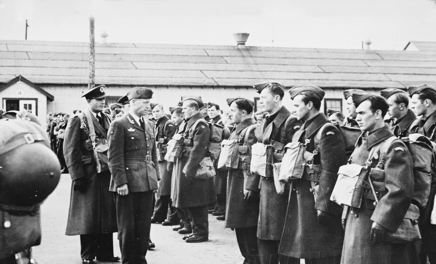 Military officers inspect soldiers standing before rows of wooden barracks.