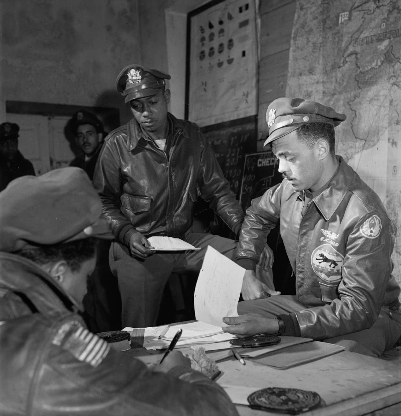 Three men in flight jackets review documents in a briefing room.